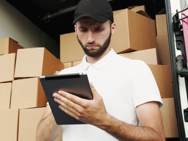 A delivery driver wearing a white polo shirt and black cap stands inside the back of a delivery truck filled with cardboard boxes, looking down at a tablet in his hands.