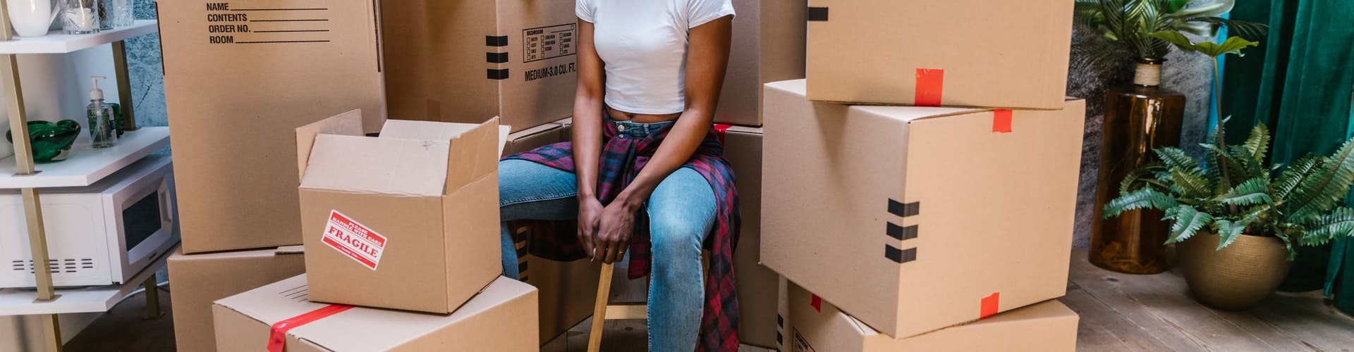A person sitting on a stool in a room surrounded by moving boxes, including one marked with a red "FRAGILE" label, indicating an active packing or unpacking process during a move.