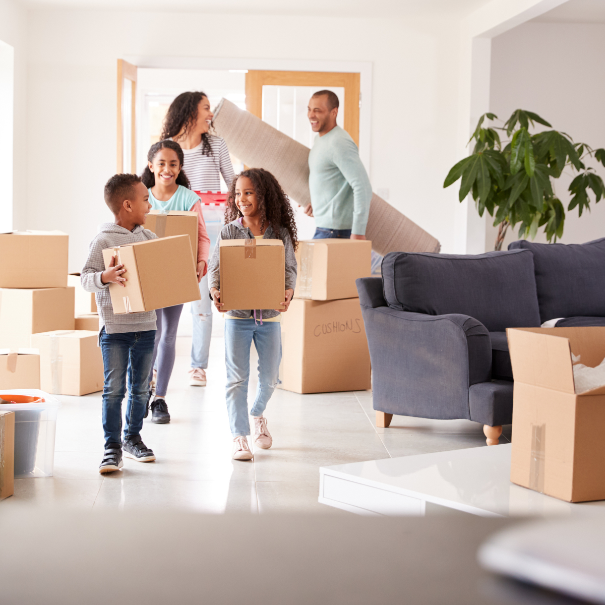A family of two adults and three children carries moving boxes and furniture into a bright new home; the children lead with smaller boxes while the adults follow with a rug and box.