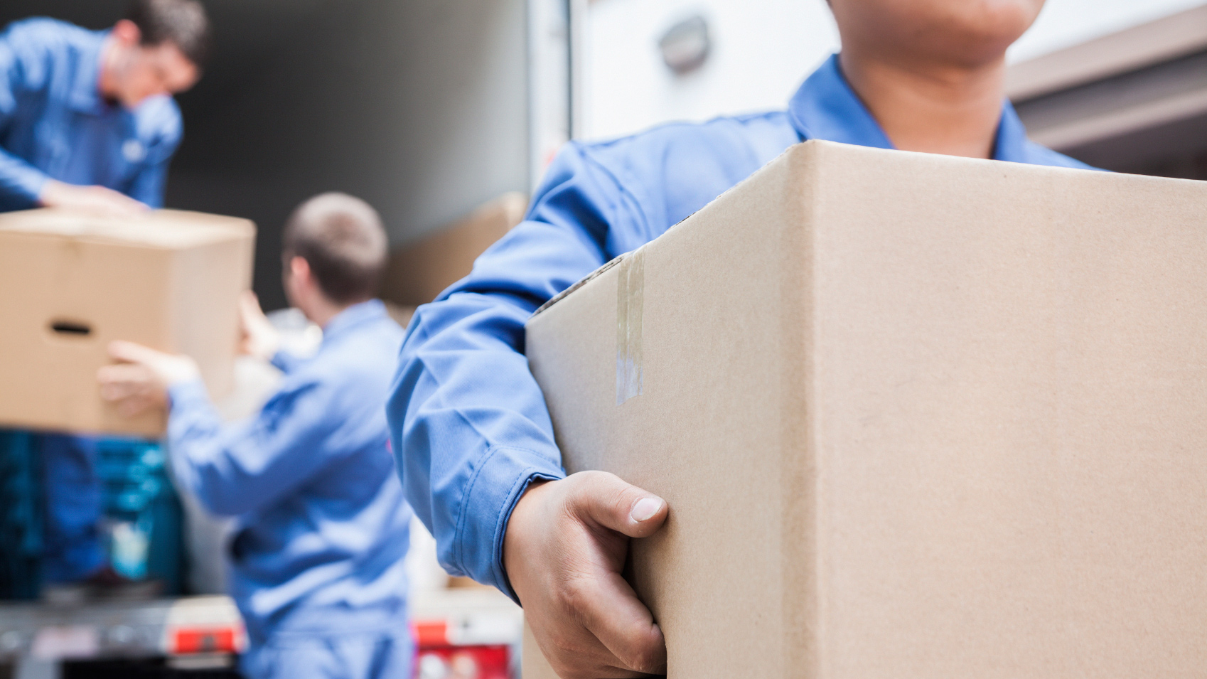 Three movers in blue uniforms load boxes onto a truck; one carries a large box in the foreground while two work inside the truck.