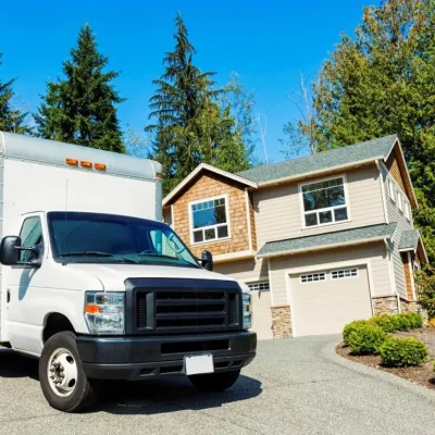 A white moving truck is parked on a driveway in front of a two-story house, with another house and tall evergreens nearby under a clear blue sky.
