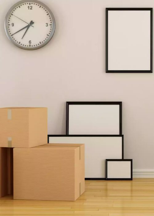 A room being prepared for moving, with a stack of cardboard moving boxes, several empty picture frames leaning against a wall, a wall clock, a white radiator, and a green ladder positioned nearby. The space has light-colored walls and wooden flooring.
